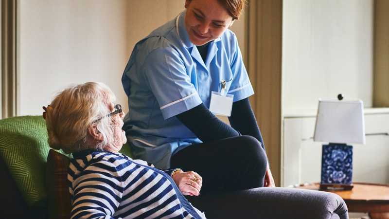 A photograph of an older women sat on a chair at home, with a health and care professioanl in a light-blue uniform sitting on the arm rest of the chair. The two women are looking at one another and smiling.