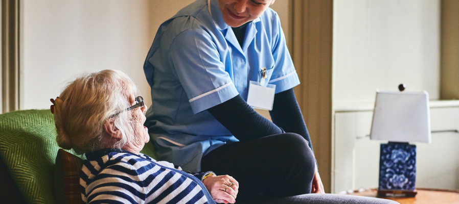 A photograph of an older women sat on a chair at home, with a health and care professioanl in a light-blue uniform sitting on the arm rest of the chair. The two women are looking at one another and smiling.