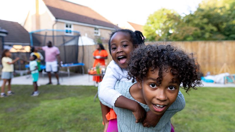 A photograph showing two children in a back garden. In the foreground a child is giving a little girl a piggy back. In the background adults and children are talking and playing next to a trampoline.