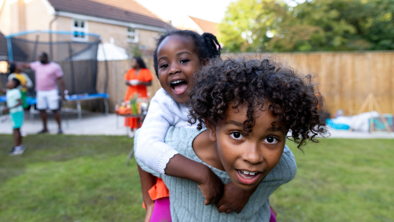 Two smiling children playing outside, one carrying the other on their back, with adults and a trampoline visible in the background.