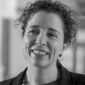 Smiling woman with curly hair, wearing earrings and a necklace, in a black and white close-up portrait.