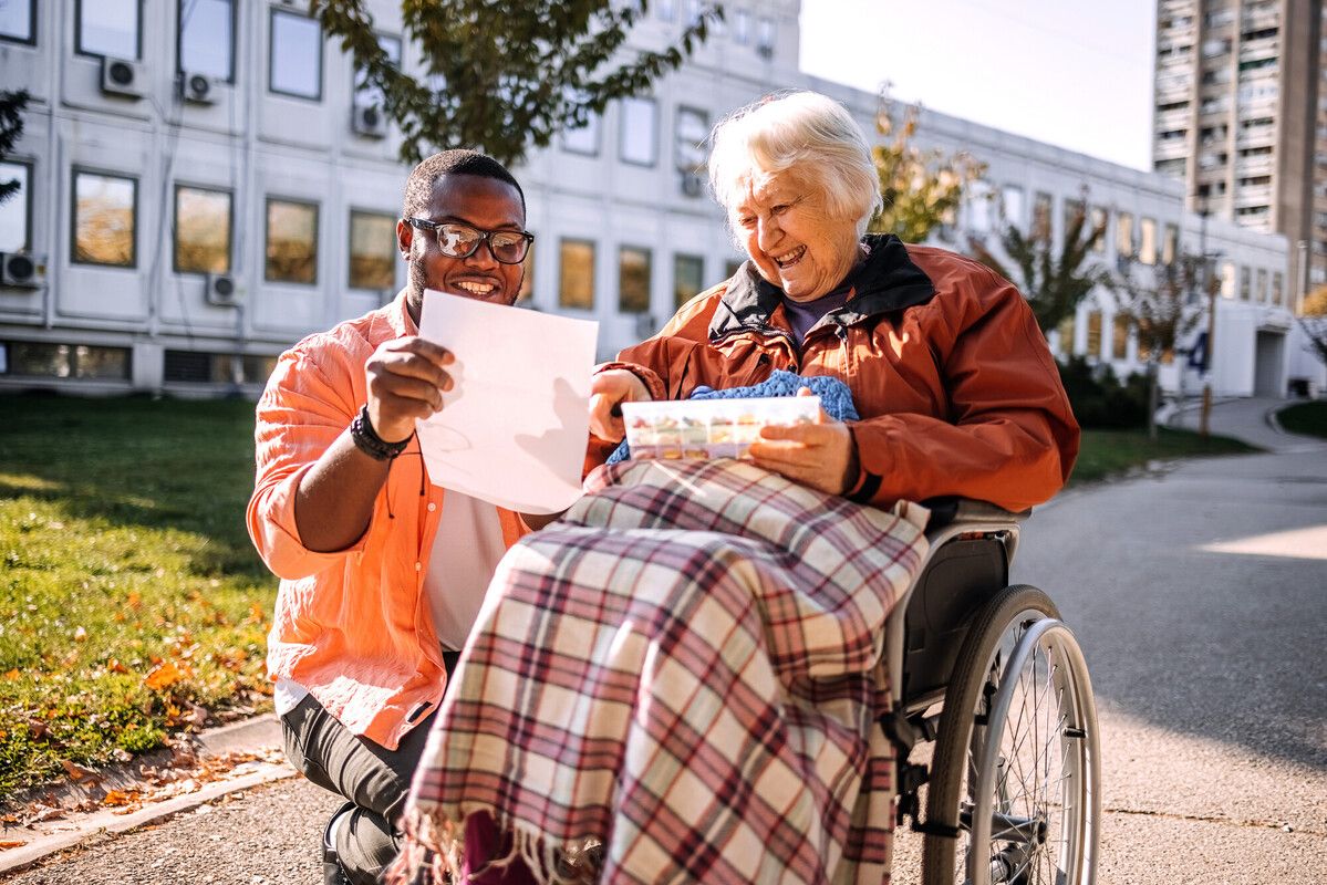 A man and an elderly woman in a wheelchair share a joyful moment outdoors, looking at a paper together, with a plaid blanket on her lap.