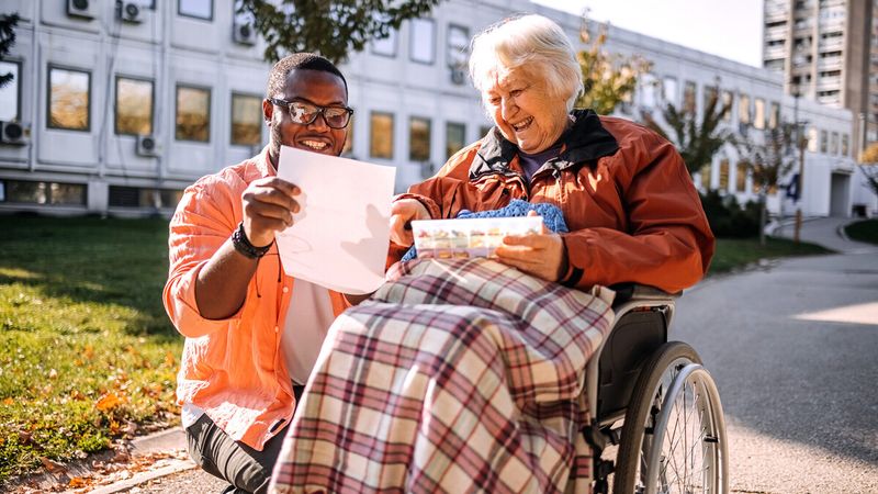 A man and an elderly woman in a wheelchair share a joyful moment outdoors, looking at a paper together, with a plaid blanket on her lap.
