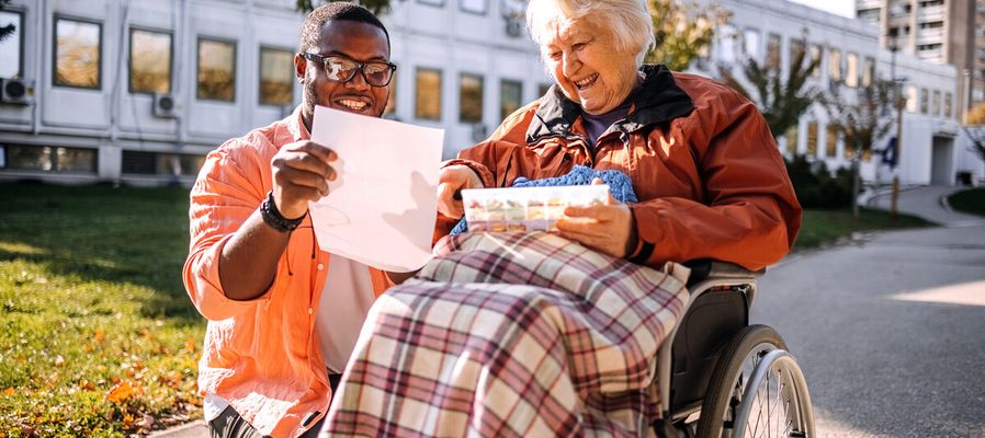 A man and an elderly woman in a wheelchair share a joyful moment outdoors, looking at a paper together, with a plaid blanket on her lap.