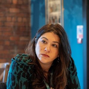 Woman sitting at a table with a notepad, looking to the side, in a room with brick walls and framed pictures.