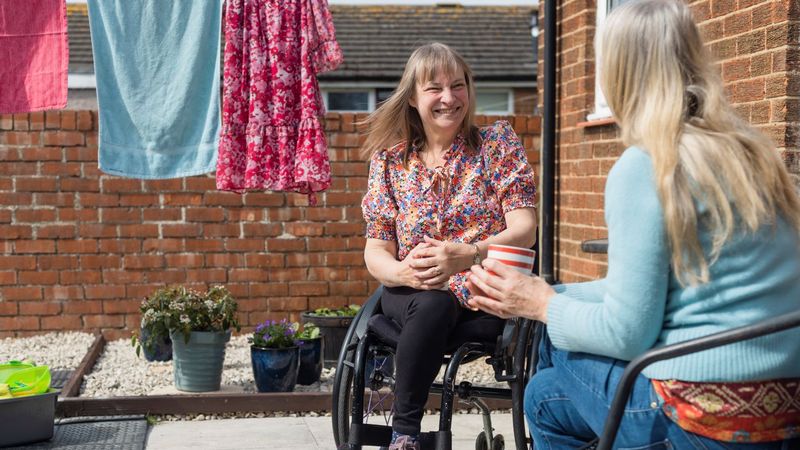 Two women talking outdoors in a back garden. One woman is in a wheelchair, and they are surrounded by hanging clothes and potted plants.
