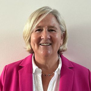 Smiling person with short blonde hair wearing a pink blazer and white shirt against a plain background.