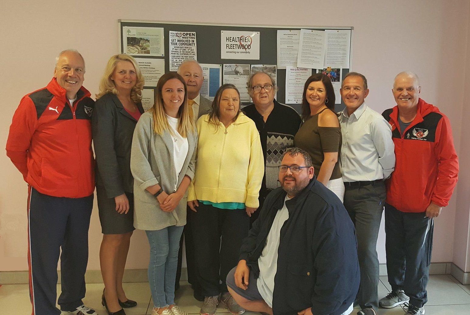 A group of eleven people smiling in front of a bulletin board displaying community information.