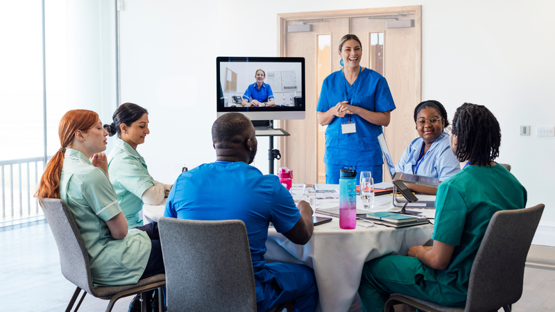 A group of medical professionals in a hybrid meeting including someone calling in on a screen.