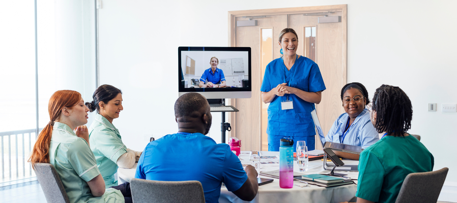 A group of medical professionals in a hybrid meeting including someone calling in on a screen.