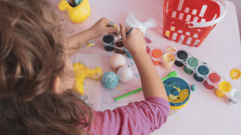 Child painting on a table, surrounded by paint pots and a red basket.