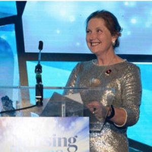 A woman in a shiny silver dress stands at a podium, smiling, with a microphone in front of her against a blue-lit backdrop.