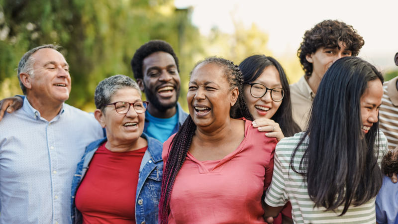 A photograph of a group of a diverse group of people, of different ages and ethnicities, outside. The people in the photo are stood closely together and laughing.