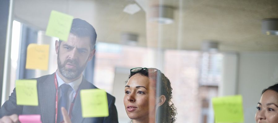 Healthcare professionals in discussion, examining colourful sticky notes on a glass board, one holding a notepad, another pointing.