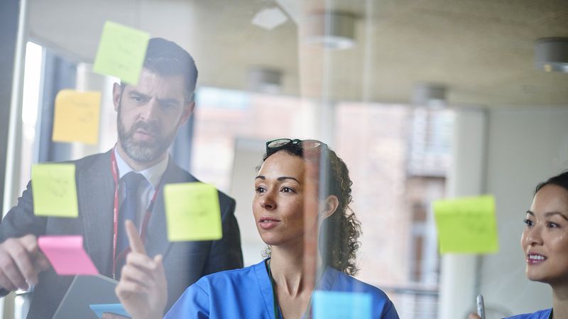 Three professionals, including two in scrubs, collaborate around a glass wall covered with colourful sticky notes in a heathcare setting.