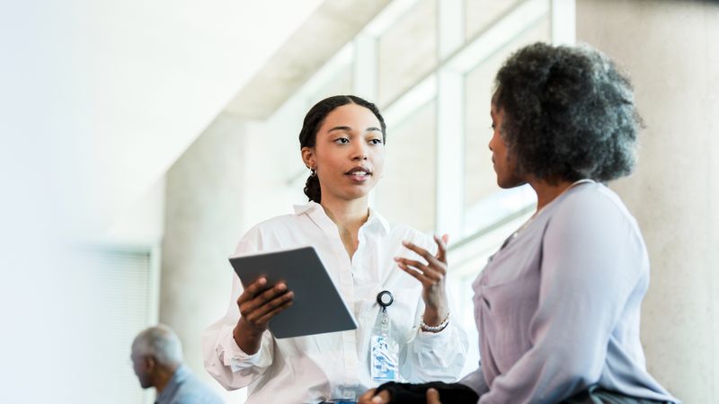 Two women are speaking. One is holding a tablet and addressing the other.