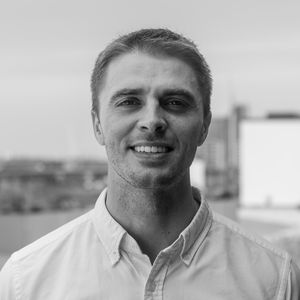 Smiling man in a white shirt standing outdoors with a blurred cityscape in the background, in a black and white photograph.