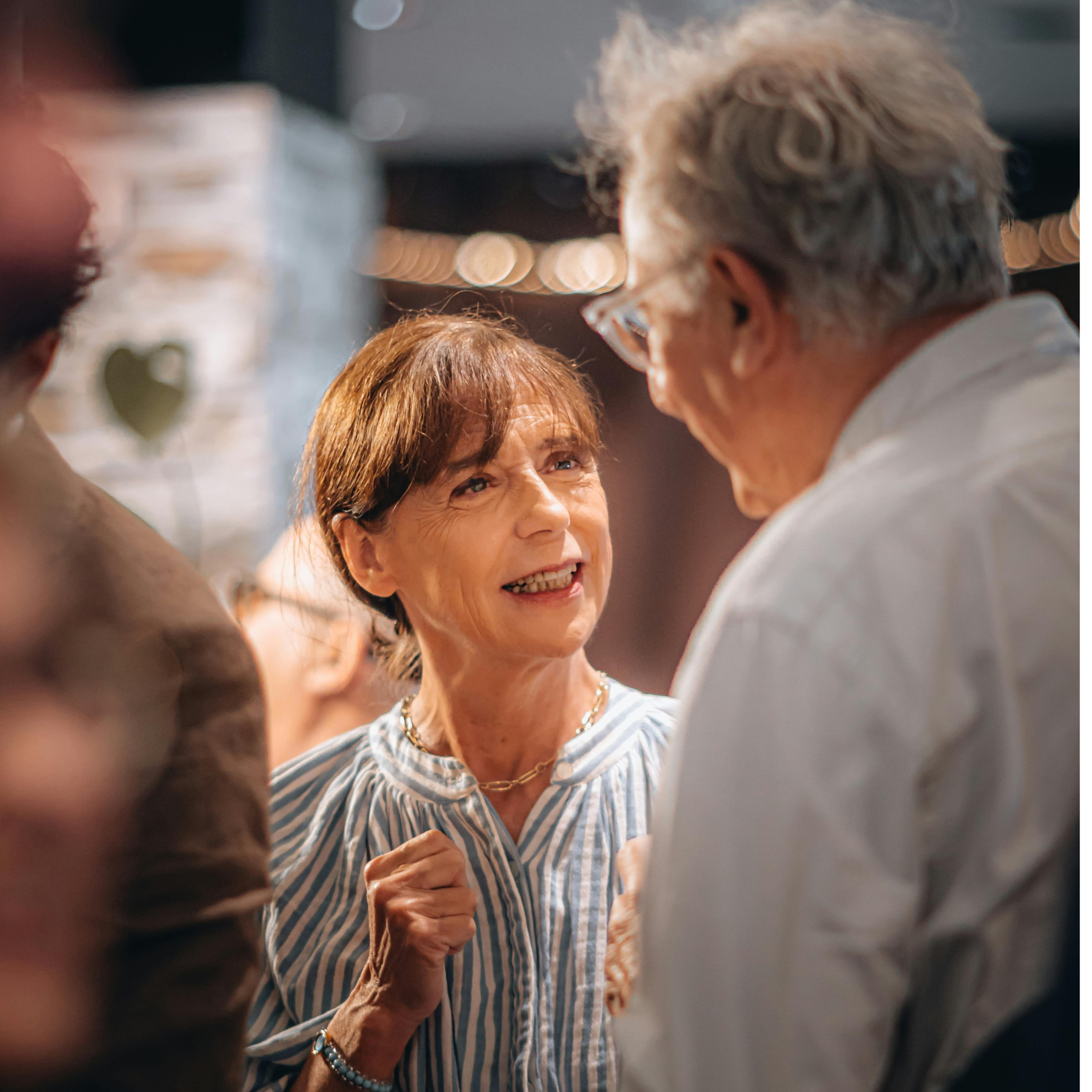 A woman in a striped shirt talks animatedly with an older man in glasses, surrounded by softly blurred lights and people in the background.