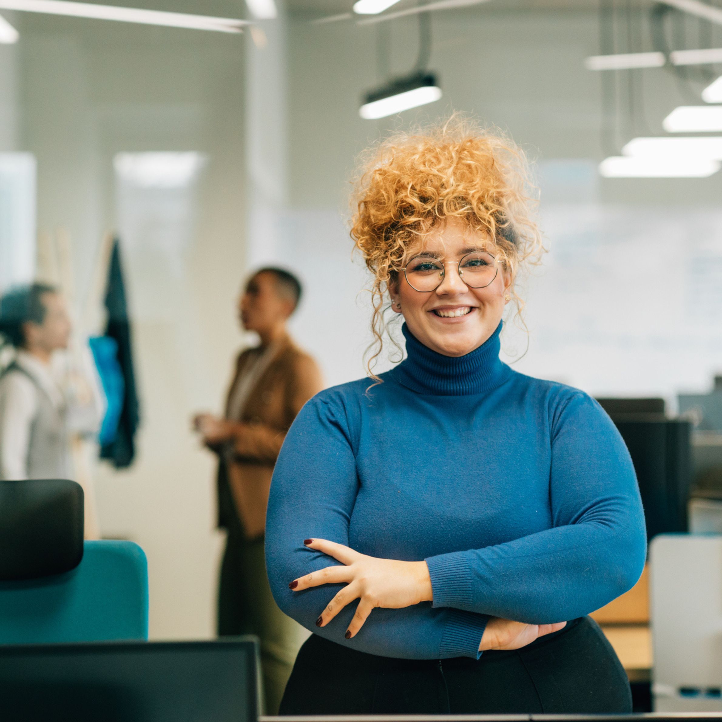 Smiling woman with curly hair and glasses in a blue turtleneck, standing with arms crossed in an office setting with colleagues in the background.