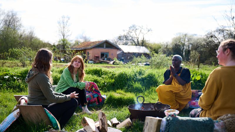Four people sitting around a fire pit in a grassy field, engaging in conversation. A house and trees are in the background.