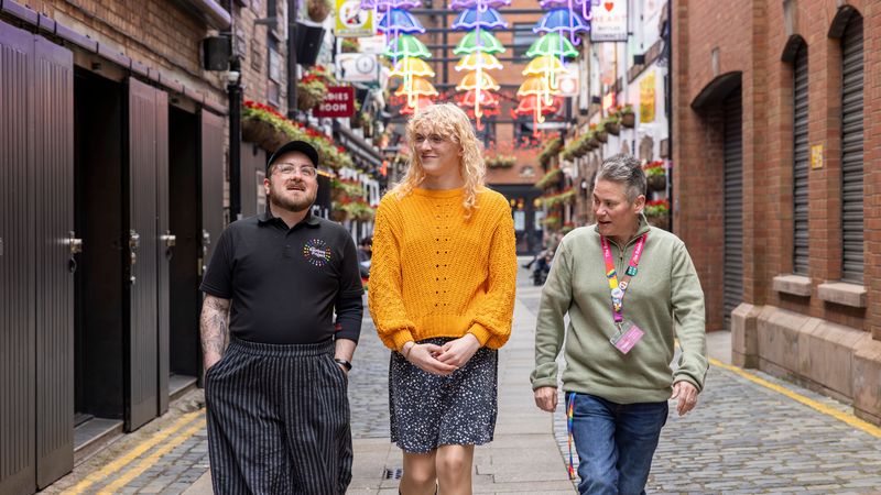 Three people walking in a colorful, neon-lit alleyway with hanging flower baskets, engaged in conversation.