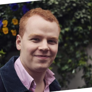 Smiling person with red hair in a pink shirt and dark jacket, standing outdoors against a leafy backdrop with yellow and purple flowers.