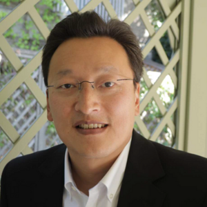 A man wearing glasses and a black suit jacket over a white shirt, standing in front of a lattice with greenery in the background.