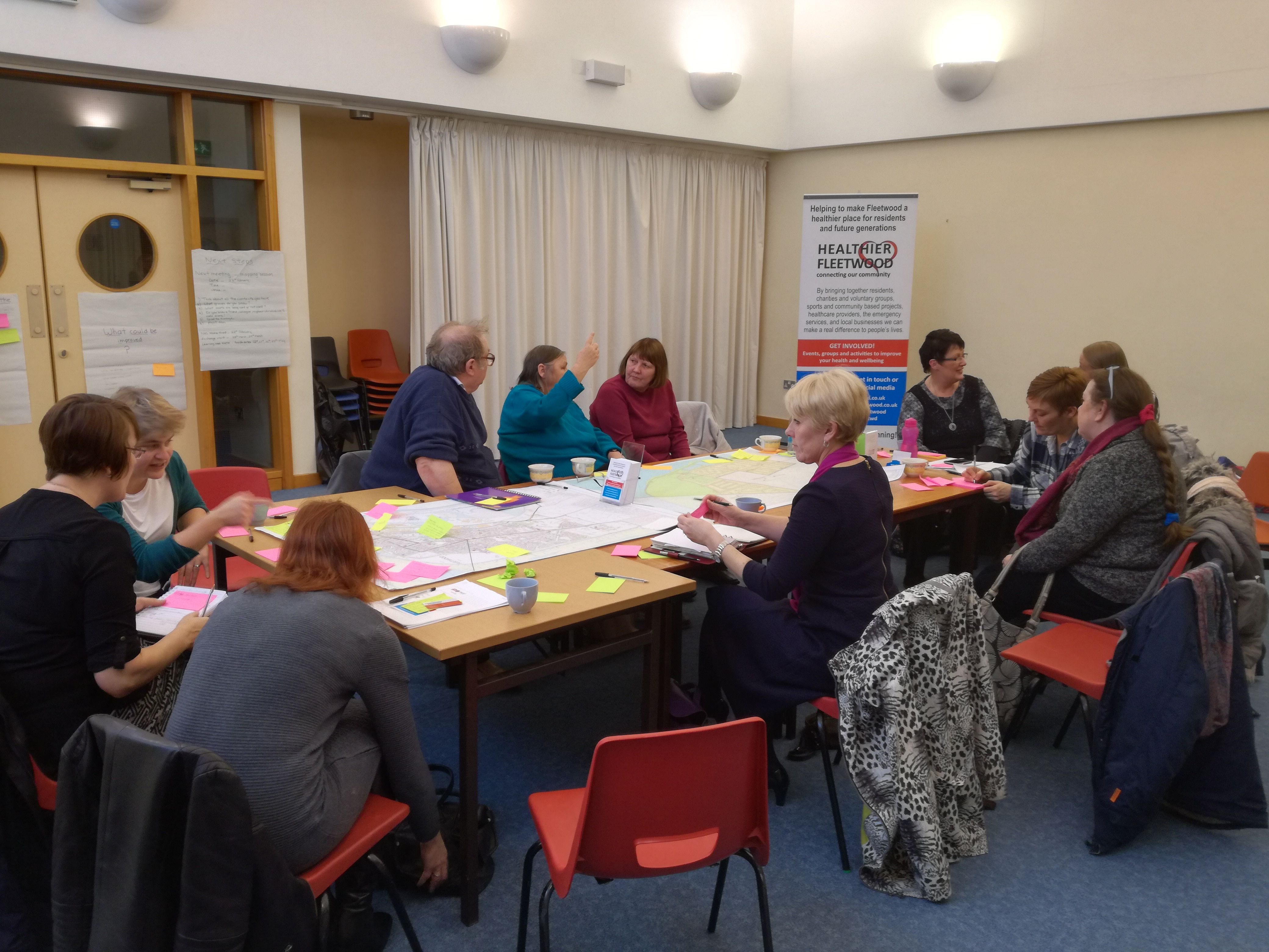 A group of people in a meeting room collaborate at tables with papers and colorful sticky notes. A 