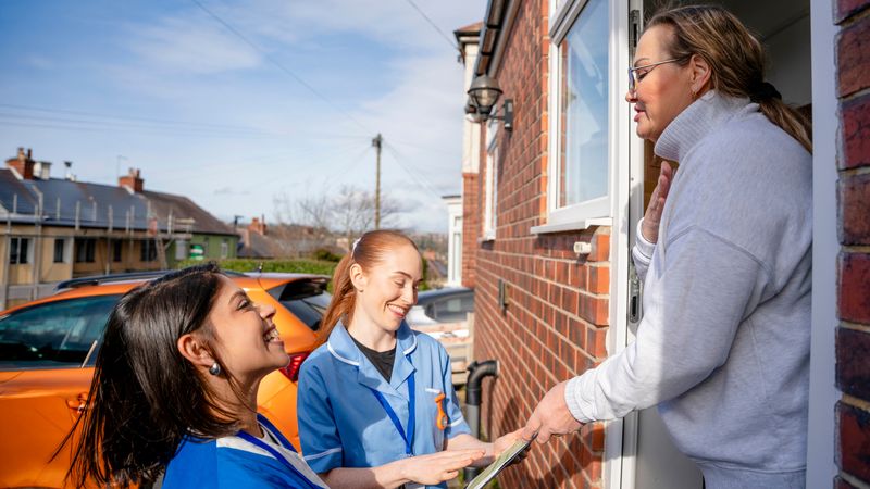 Two healthcare workers in blue uniforms smile while talking to a woman at her doorstep on a sunny day.