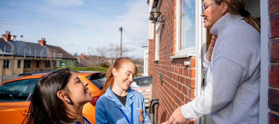 Two healthcare workers in blue uniforms smile while talking to a woman at her doorstep on a sunny day.