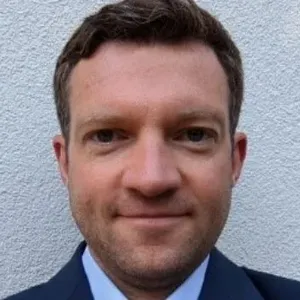 A man with short brown hair wearing a dark suit and tie, standing in front of a textured white wall, looking directly at the camera.