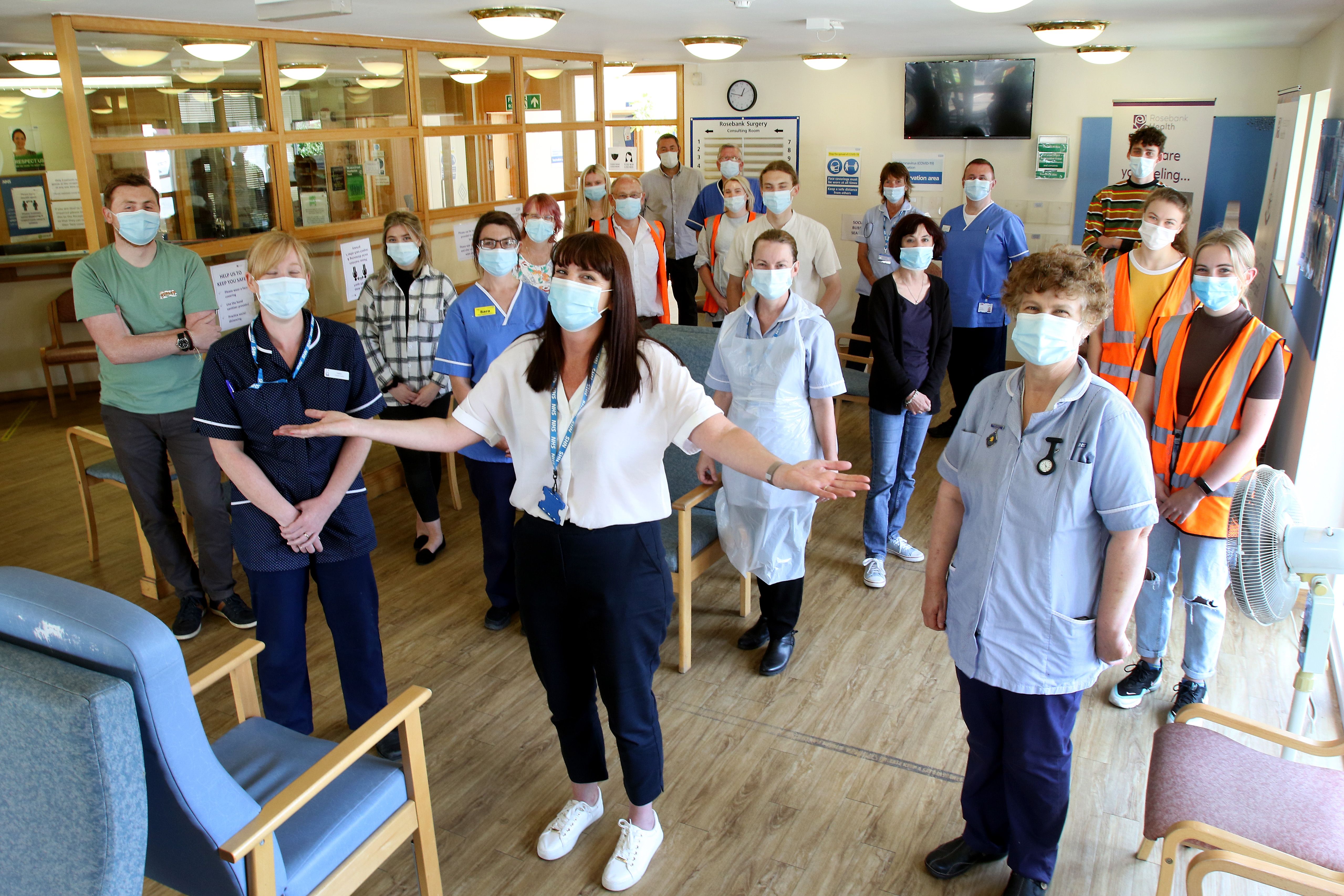 Masked medical staff and volunteers pose in a clinic waiting room, a woman in front gesturing with her arms outstretched.