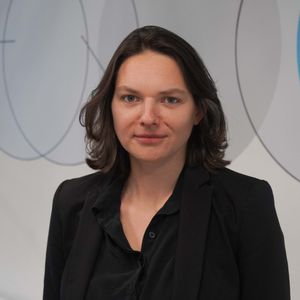 Woman with shoulder-length dark hair wearing a black blazer, standing in front of a white wall with geometric blue and gray patterns.