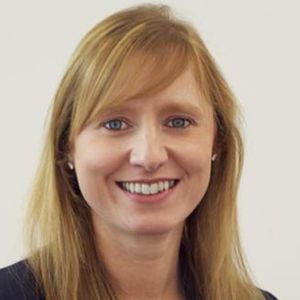 Smiling woman with long light brown hair wearing a dark blazer against a light background.