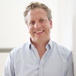 Smiling man with curly hair in a light blue checkered shirt standing against a white wall.