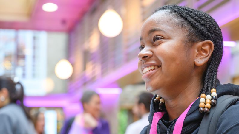 A person with braided hair and bead accessories smiles warmly in a modern space with pink lighting and blurred people in the background.