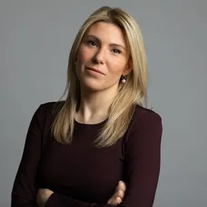 Woman with long blonde hair in a dark top stands confidently with arms crossed against a gray background.