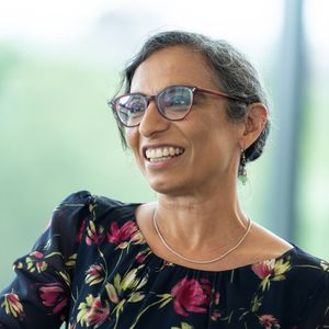 Smiling woman with glasses and floral dress in a bright, blurred background setting.