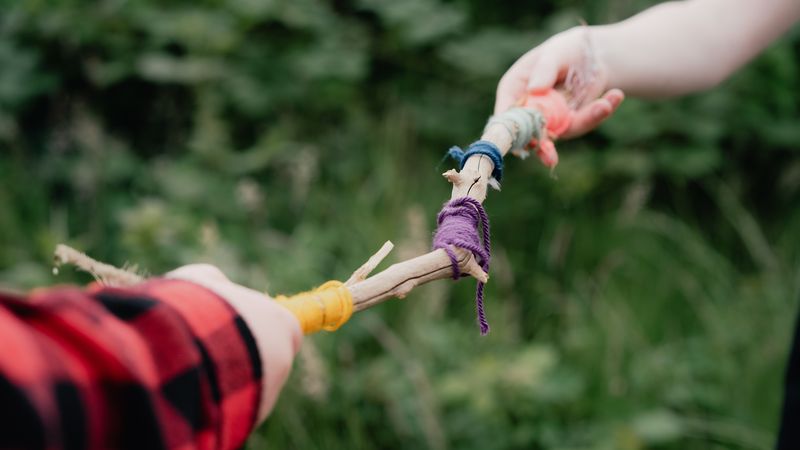 Two hands holding a stick wrapped with colourful yarn, against a blurred green foliage background.
